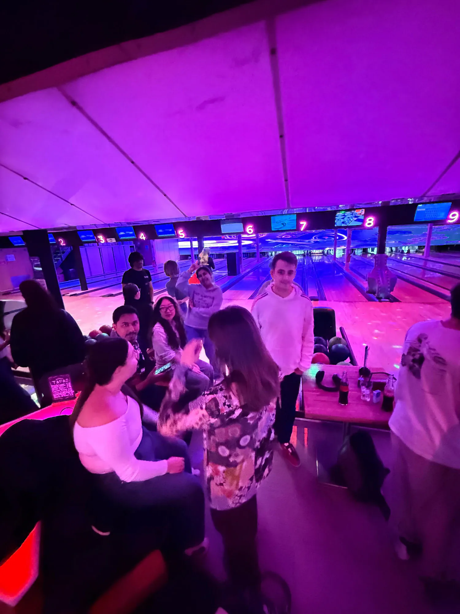 A group of students gathered around a bowling lane under purple neon light, drinks and bowling balls nearby