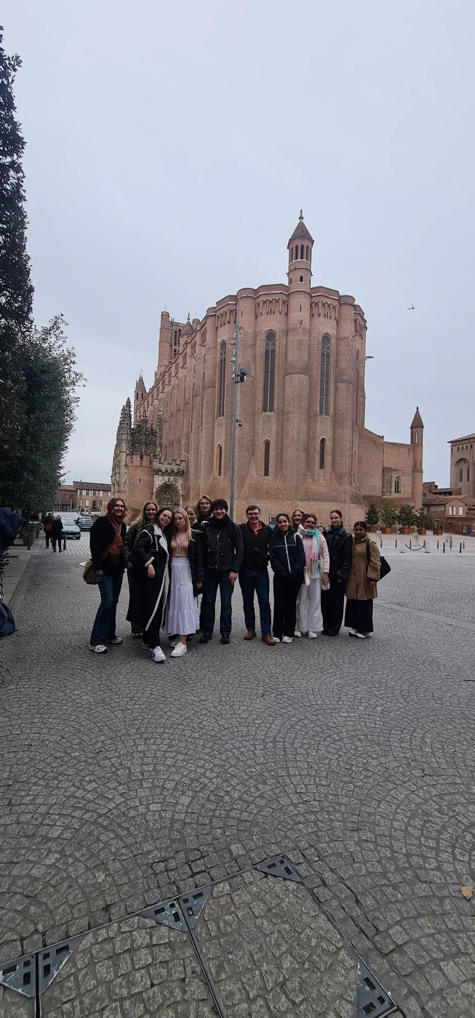 A dozen AEGEE Toulouse members posing together on the cobbled square behind the red brick Sainte-Cécile cathedral in Albi