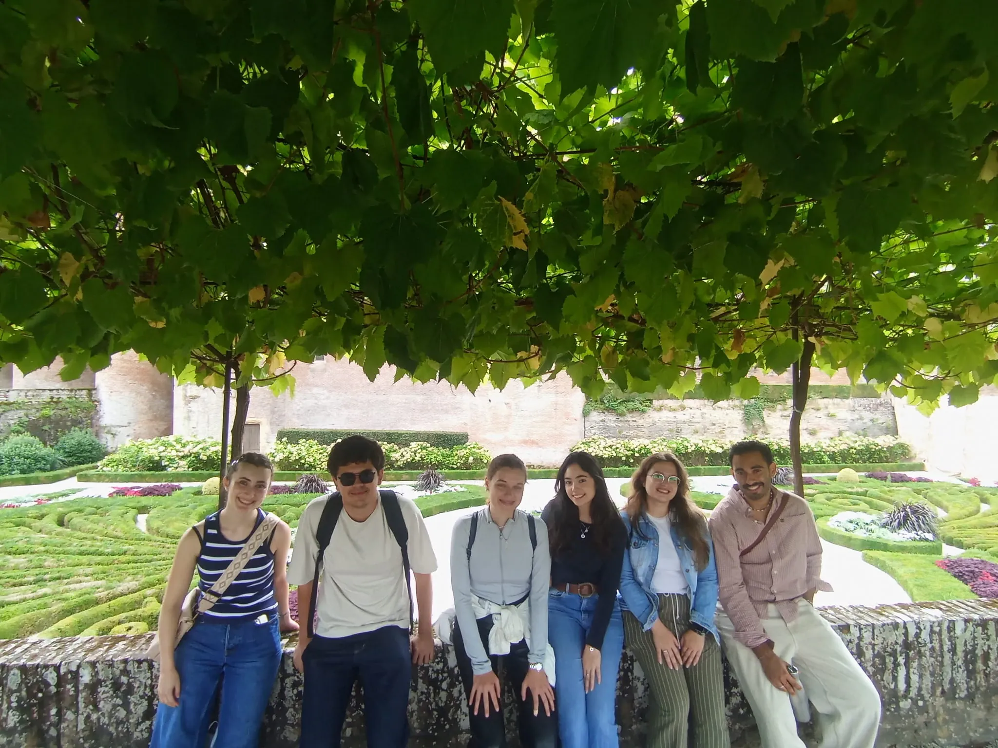 A group posing under a grape vine trellis in a formal garden, clipped hedges behind them