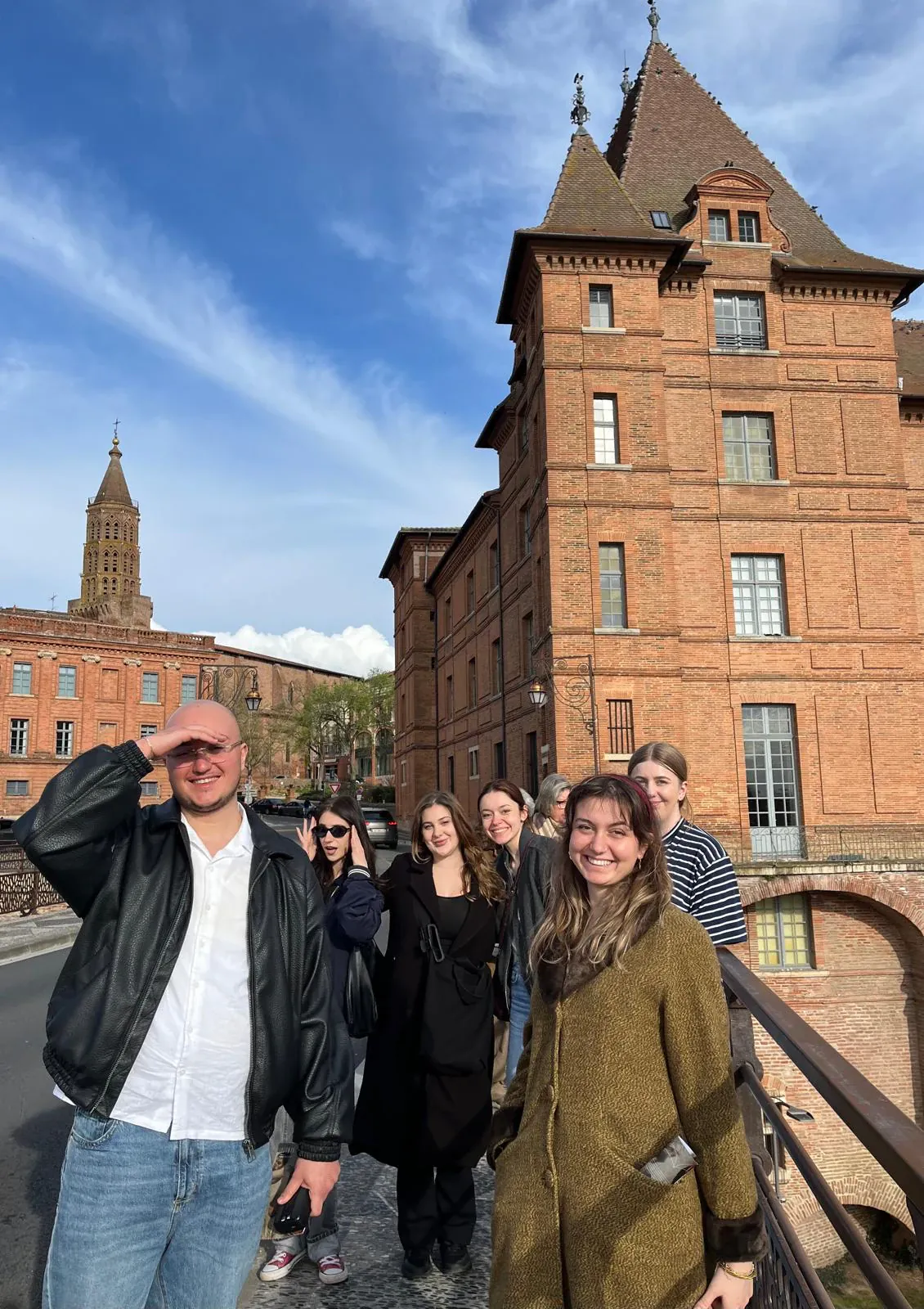 A group standing on a bridge in front of a tall red-brick building under a blue sky