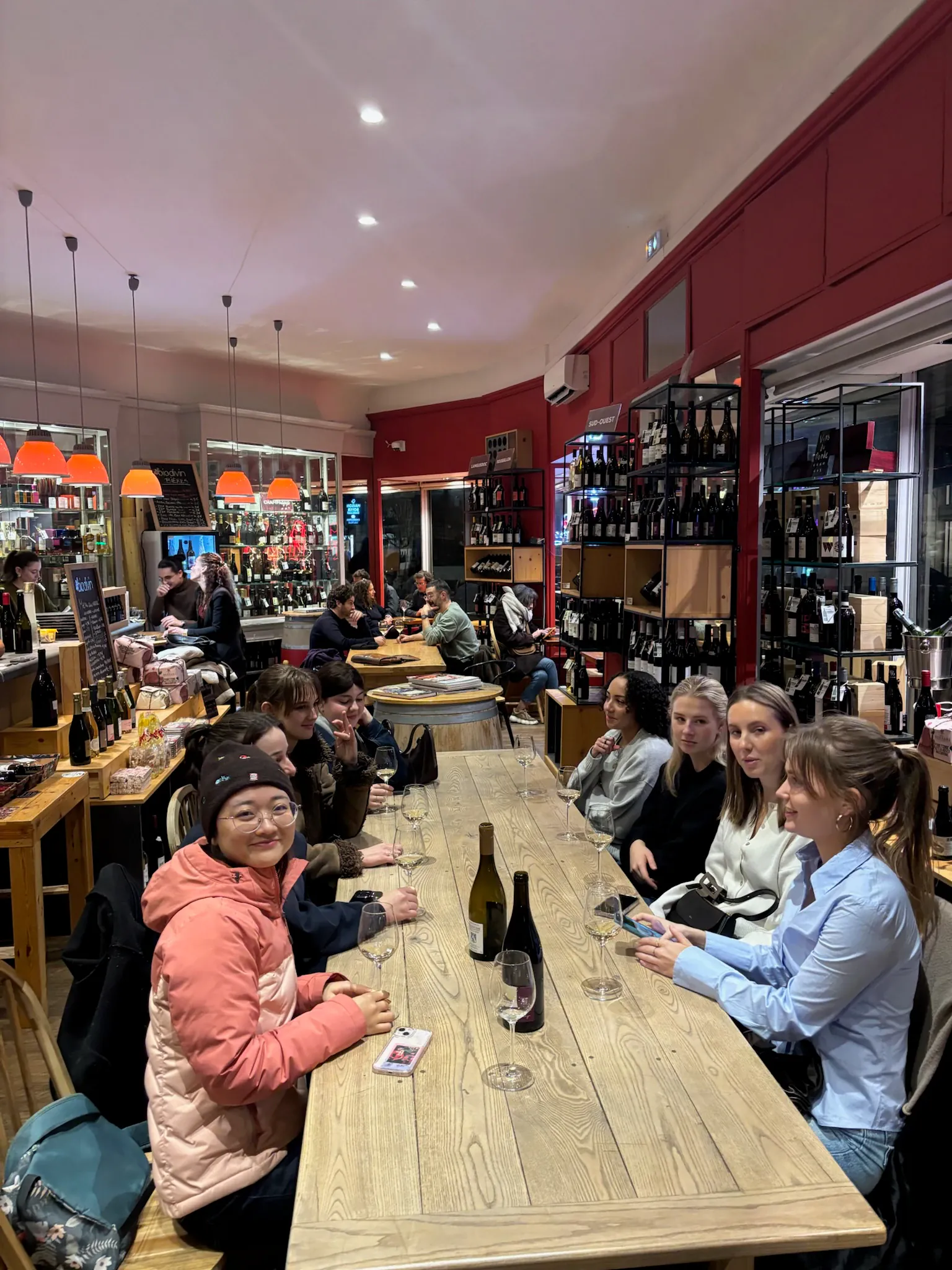 A group seated around a long wooden table in a wine shop, glasses of white wine in front of them