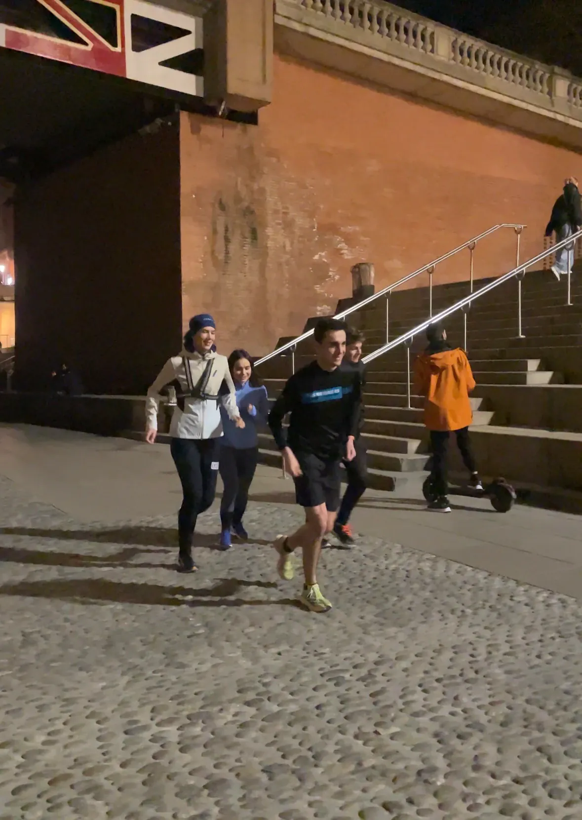 A group of runners smiling mid-stride on a cobbled riverside path in Toulouse at night
