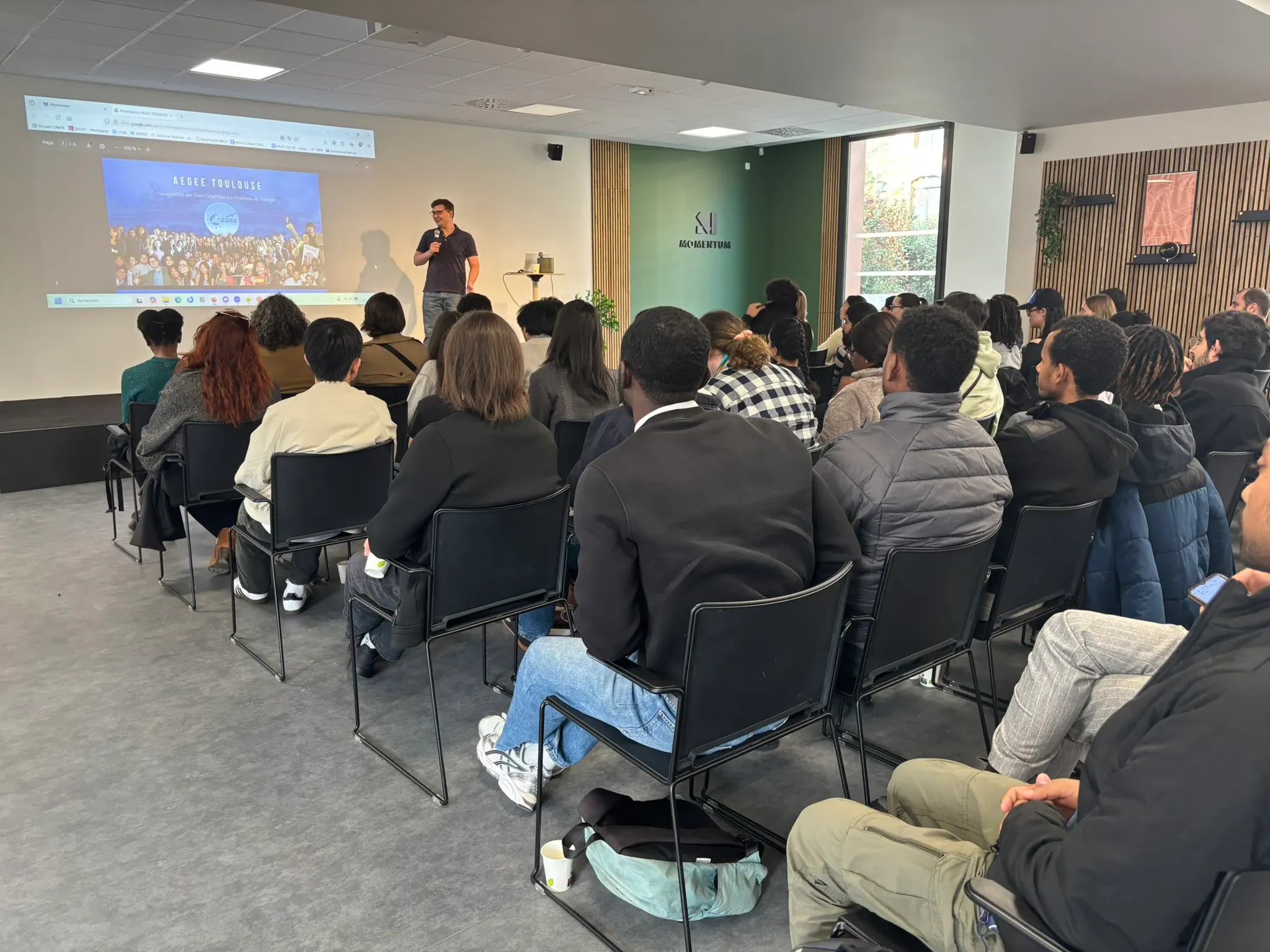 A member presenting to a full room of seated students, a title slide projected behind them