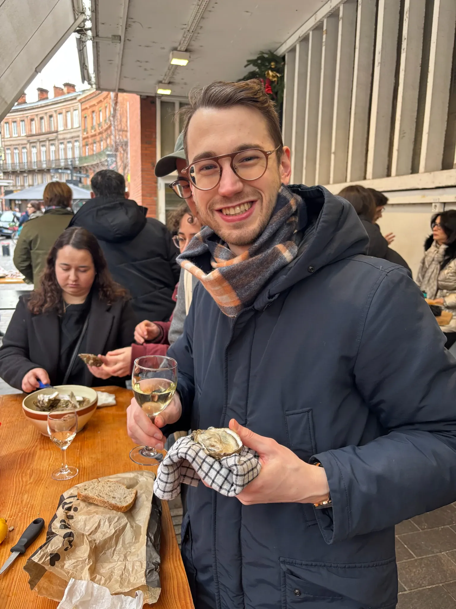 A student smiling while holding an open oyster and a glass of white wine outdoors