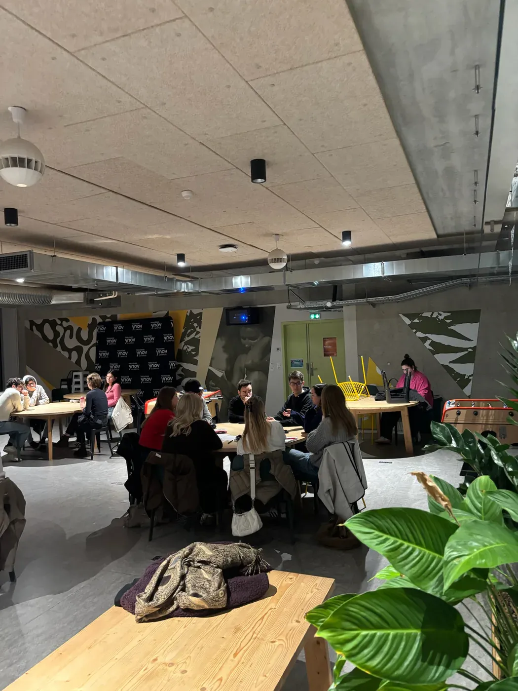 A campus lounge with students grouped around long wooden tables, a painted wall behind them