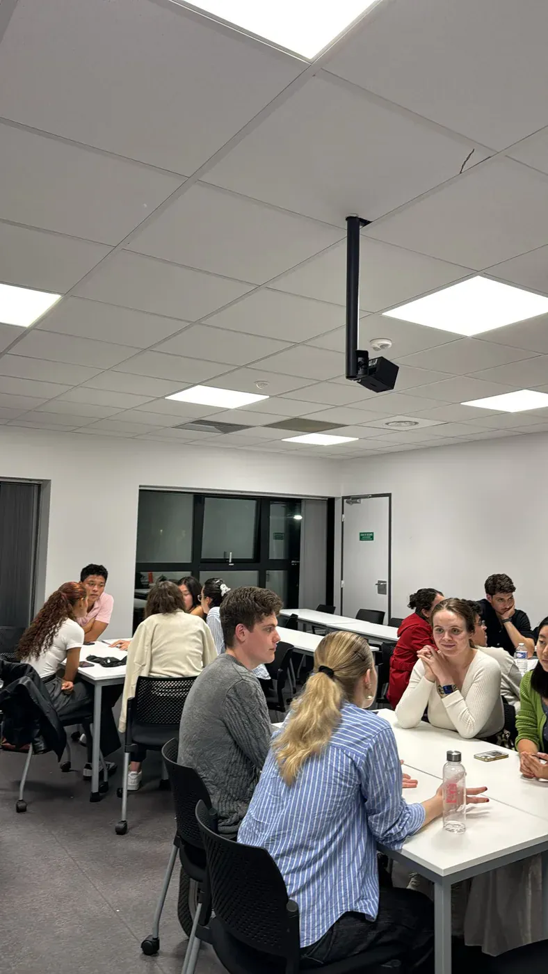 Students seated in twos and threes around classroom tables, talking to each other during a language workshop