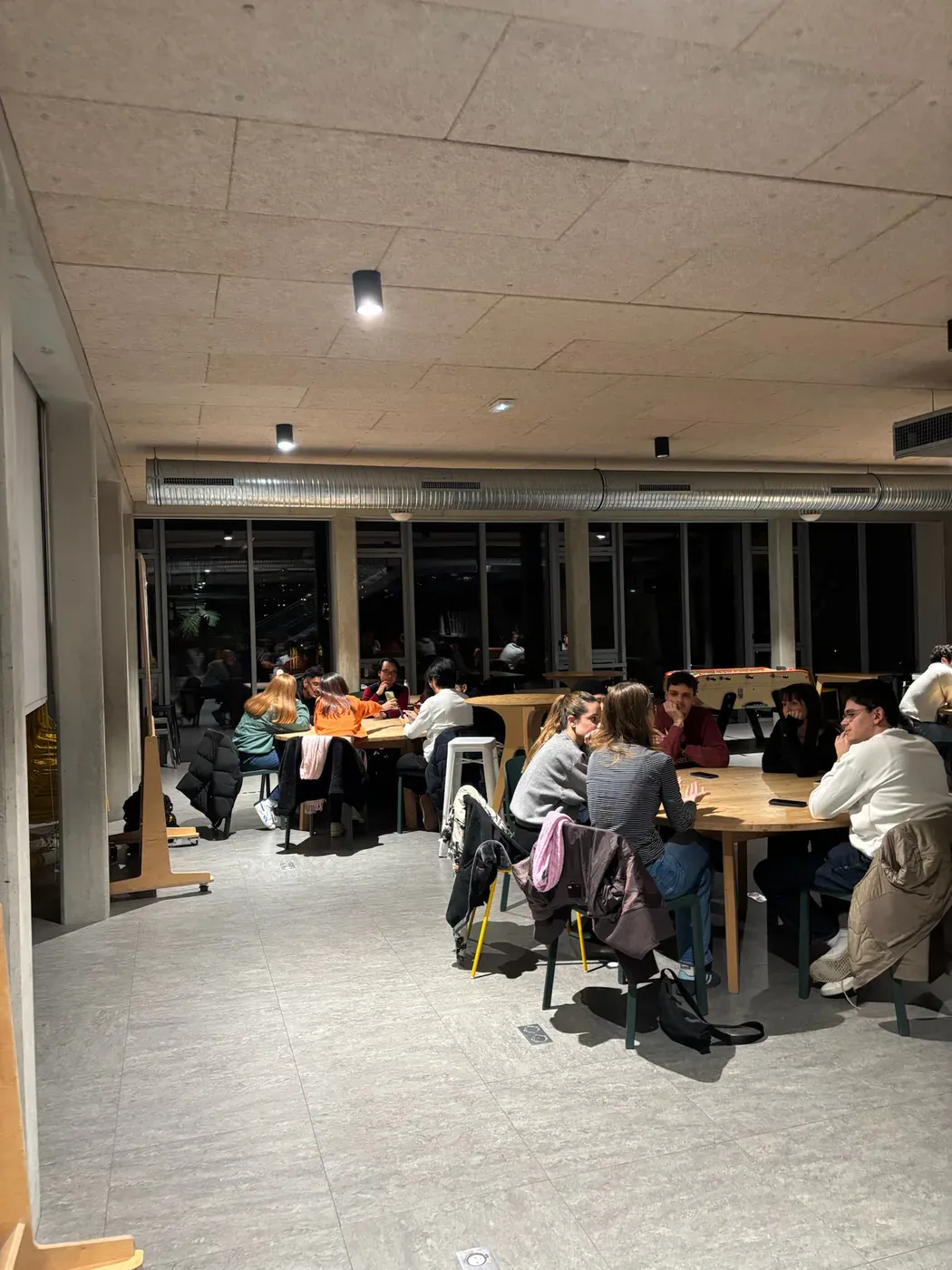 A campus room in the evening with students seated at tables along a wall of dark windows