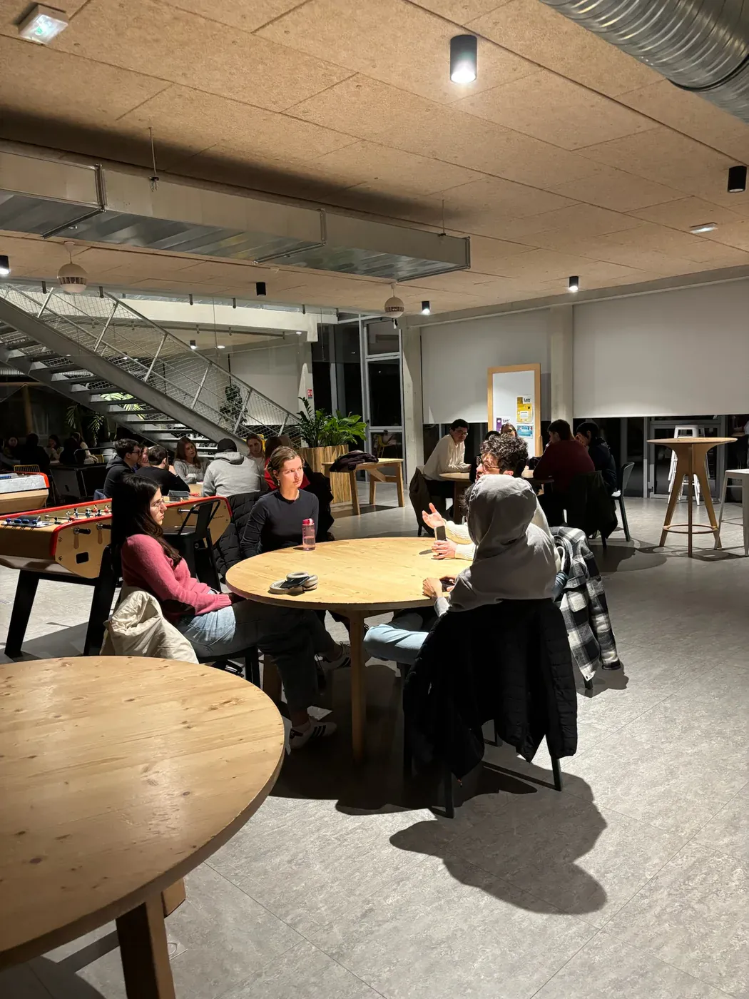 Two round tables of students in conversation in a campus common room, a table football in the background